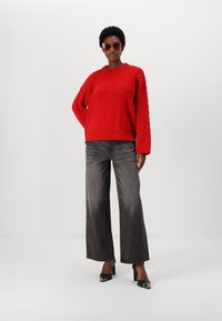 Raudonas megztinis su plataus pynimo rankovėmis, derinamas su aukštu juosmeniu, platėjančiomis pilkomis džinsomis ir juodais smailianosiais aukštakulniais.