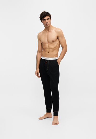 Homme torse nu aux cheveux foncés, debout pieds nus, portant un pantalon de jogging noir avec une ceinture élastique blanche sur un fond clair uni.