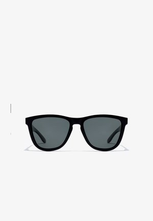 Gafas de sol negras con forma rectangular y lentes oscuras, montura mate texturizada, combinadas con un suéter marrón de punto. Accesorios incluyen un pendiente de cruz plateada.