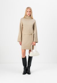 Beige stickad klänning med hög krage och bälte i midjan, kompletterad med en vit handväska och svarta knästövlar. Slät textur, avslappnad passform.