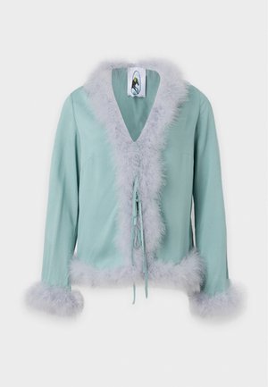 Un mannequin porte une tenue deux pièces bleu sarcelle pâle avec une bordure en fourrure lavande et des talons dorés, sur un fond blanc uni.