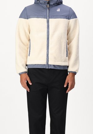 Mann trägt beige Fleecejacke mit blauen gepolsterten Schultern und Kapuze, Reißverschluss vorne und schwarze Hose, steht vor weißem Hintergrund.