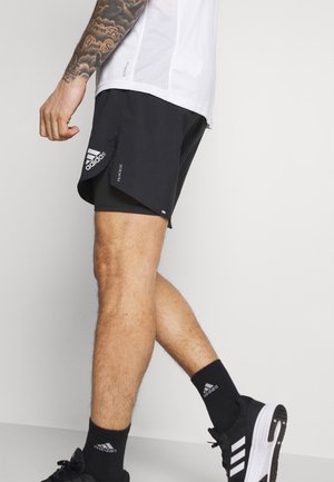 Shorts de sport noirs avec une fente sur le côté, ornés d'un logo Adidas blanc. Associés à des chaussettes et des chaussures noires, soulignant un look sportif.
