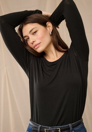 Junge Frau mit braunen Haaren, die ein schwarzes langärmeliges Shirt und blaue Jeans trägt, posiert mit erhobenen Armen hinter dem Kopf.