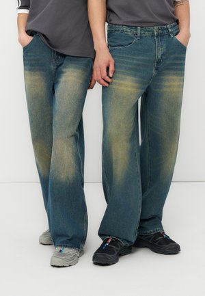 Δύο άτομα στέκονται δίπλα-δίπλα φορώντας φαρδιά ξεβαμμένα μπλε τζιν, casual γκρι και μαύρα αθλητικά παπούτσια, και σκούρα μπλουζάκια με τα χέρια στις τσέπες.