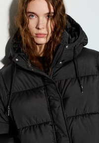 Doudoune noire avec capuche, présentant un motif matelassé, une fermeture éclair frontale et des cordons de serrage pour un ajustement réglable.