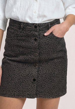 Femme portant une jupe noire et grise à pois avec boutons sur le devant, la main dans la poche et un chemisier blanc.