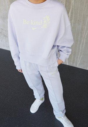 Personne portant un sweat-shirt violet clair avec le texte « Soyez gentil » et un motif floral, un pantalon assorti, et des baskets blanches, debout à l'intérieur.
