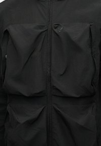 Μαύρο μπουφάν με φερμουάρ μπροστά και τέσσερις μεγάλες, ελαφρώς τσαλακωμένες τσέπες, δύο στο στήθος και δύο κοντά στη μέση, με διακριτικό κεντητό λογότυπο στις πάνω τσέπες.