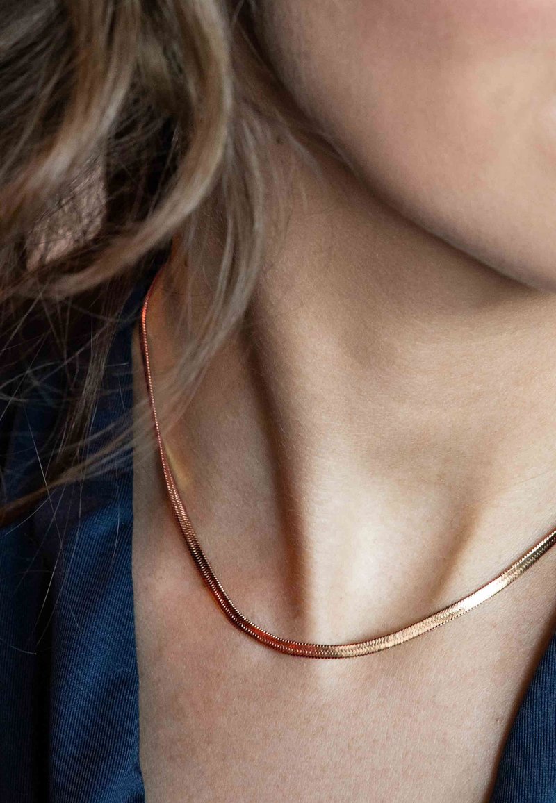 Gros plan d’un cou avec un collier fin en chaîne d’or lisse porté sur la peau et un col de chemise bleu foncé visible.