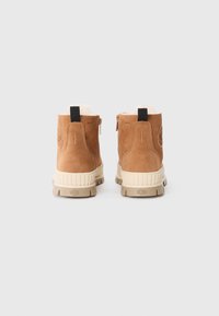 Bottines en daim marron avec une finition texturée, semelles en caoutchouc de couleur crème et doublure douce, présentant une languette noire et un détail de fermeture éclair.