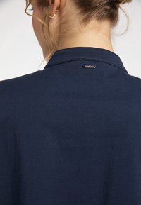 Camicia blu navy con una texture liscia, dotata di colletto e un piccolo dettaglio di logo metallico sul retro. Design pulito e minimalista.