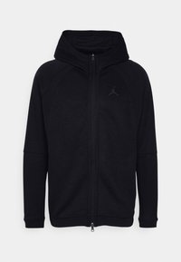 Černá mikina na zip s vysokým límcem, vyrobená z měkké tkaniny. Má raglánové rukávy a nenápadné logo na hrudi.