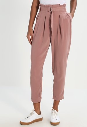 Vrouw die mauve, hoog getailleerde, taps toelopende broek met riem draagt, gecombineerd met witte sneakers, staand tegen een lichte achtergrond.