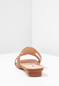Tan flat sandals featuring an open back, two wide straps, stitched accents, and a textured sole. The footbed is cushioned for comfort.
