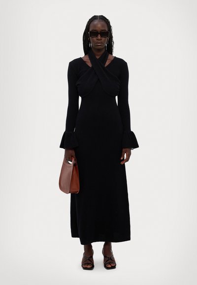 Robe noire à manches longues avec un décolleté croisé et des poignets évasés. Le mannequin tient un sac à main marron et porte des lunettes de soleil noires.