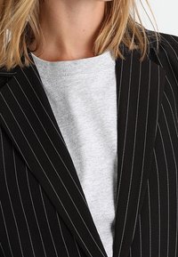 Blazer noir à fines rayures avec larges revers porté sur un sweat-shirt gris clair à col rond. Tissu texturé avec un motif vertical marqué.