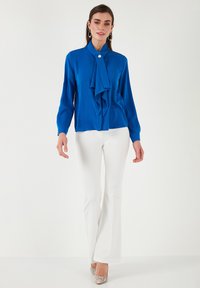 Bluză albastră cu un nod la gât, închidere cu nasturi, mânecă lungă; asortată cu pantaloni evazați albi și pantofi cu toc metalici.