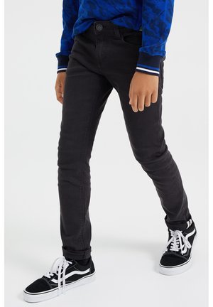 Junge trägt enge schwarze Jeans, ein blau gemustertes Langarmhemd und schwarze Sneakers mit weißen Schnürsenkeln, steht mit auseinanderstehenden Beinen.