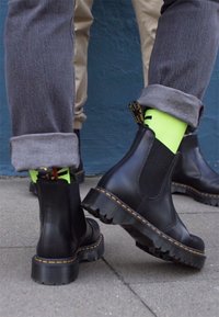 Stivali Chelsea neri in pelle con pannello laterale texturizzato e cuciture gialle; indossati con pantaloni grigi piegati e calze neon verdi vivaci.