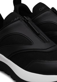 Baskets slip-on noires Calvin Klein avec fermeture éclair latérale et semelle blanche texturée, présentant un mix de panneaux en mesh et synthétiques.