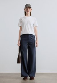 Λευκό βαμβακερό t-shirt με κεντημένο λογότυπο, συνδυασμένο με μπλε σκούρο παντελόνι φαρδιάς γραμμής. Καφέ δερμάτινη τσάντα ώμου και γκρι καπέλο με επίπεδο γείσο.