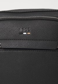 Borsa in pelle nera testurizzata con chiusura a zip; presenta il logo del marchio impresso e tre accenti di colore: nero, bianco e oro.