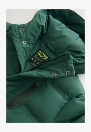 Žalias nepralaidus striukės pamušalas, pasižymintis lygiu tekstūru, su užtrauktuku užsegama kišenė ir logotipo etiketė su priežiūros instrukcijomis.