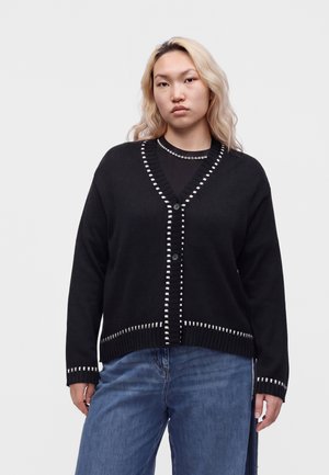Junge Frau mit langen blonden Haaren trägt eine schwarze Strickjacke mit weißen Nähten über einem schwarzen Top und blauen Jeans vor einem einfarbigen Hintergrund.