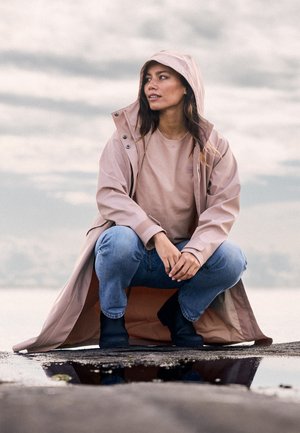 Kobieta przykucnięta na mokrej ziemi, ubrana w jasnoróżową kurtkę przeciwdeszczową z kapturem, dżinsy i czarne buty, spoglądająca w bok pod pochmurnym niebem.