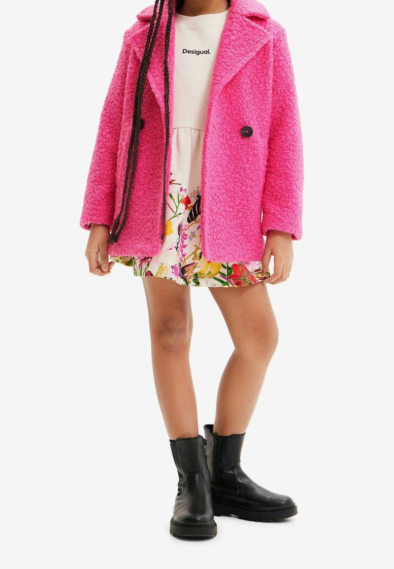 Manteau en peluche rose avec un col, boutons noirs ; porté sur une robe fleurie. Bottines noires à semelle épaisse et fermeture éclair latérale.