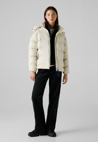 Cremfarbener Puffer-Jacke mit hohem Kragen, Reißverschluss und blauen Akzenten in lockerem Schnitt. Kombiniert mit schwarzen weit geschnittenen Hosen und dunklen Schuhen.