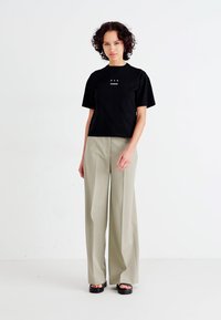 Zwarte katoenen t-shirt met korte mouwen en witte tekstlogo, gecombineerd met lichtbeige wijde broek en zwarte sandalen. Effen achtergrond.