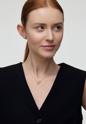 Złoty naszyjnik z przeplatanymi pierścionkami, delikatny łańcuszek, noszony przez modelkę w czarnej bluzce. Gładka tekstura i minimalistyczny design.