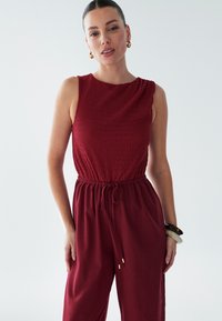 Rdeč brezrokavnik z strukturiranim zgornjim delom, elastičnim pasom in širokimi hlačnicami. Ima vrvico za zategovanje in zlate kovinske dodatke.