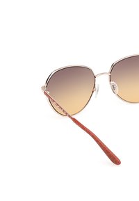 Gafas de sol de estilo gato con lentes graduales en tono marrón, marco metálico en oro rosa y patillas texturizadas en marrón rojizo que presentan un detalle de logo.