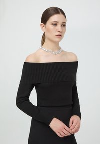 Czarna dzianinowa bluzka odkrywająca ramiona z prążkowanym dekoltem, długimi rękawami i dopasowanym krojem, zestawiona z błyszczącym naszyjnikiem z przezroczystych kamieni szlachetnych.
