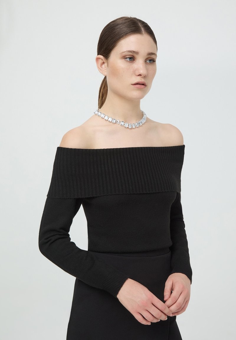 Czarna dzianinowa bluzka odkrywająca ramiona z prążkowanym dekoltem, długimi rękawami i dopasowanym krojem, zestawiona z błyszczącym naszyjnikiem z przezroczystych kamieni szlachetnych.