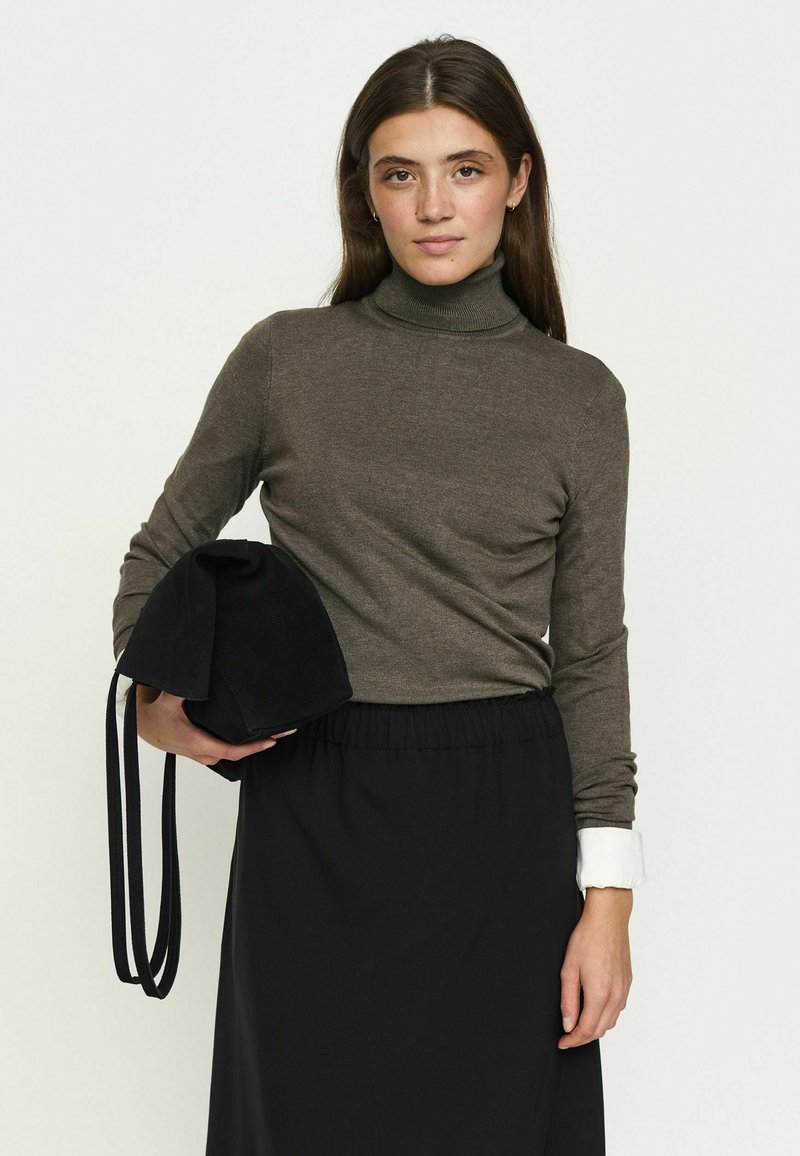 Femme aux longs cheveux bruns portant un col roulé vert olive et une jupe noire, tenant un sac noir devant un fond clair uni.