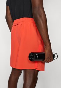 Person som bär klara orangea träningsshorts och håller en svart Nike-vattenflaska vid sin sida mot en neutral bakgrund.