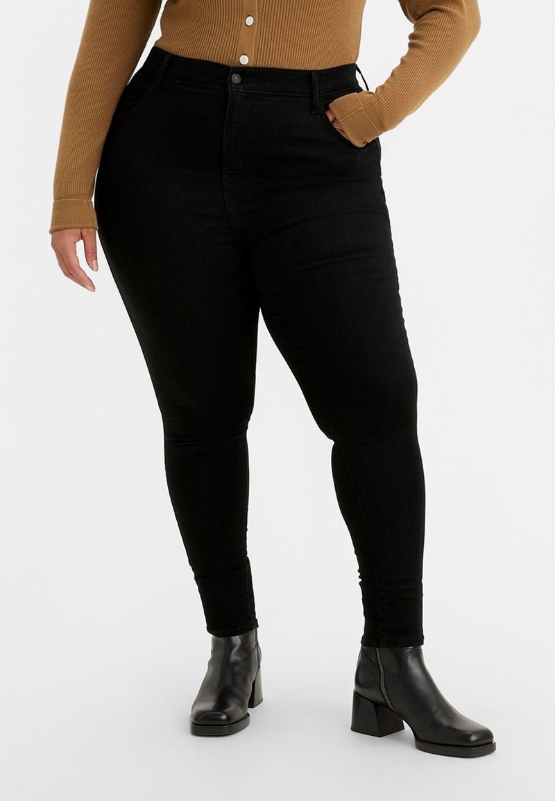 Personne portant un jean skinny noir taille haute, un pull marron boutonné et des bottines noires, debout sur un fond blanc.