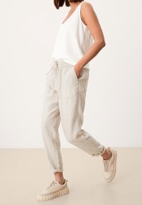 Person som bär beiga linnebyxor med dragsko, vit ärmlös topp och beiga sneakers med tjock sula, stående med ett ben lyft.