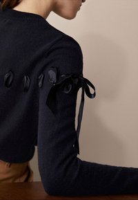 Laivastonsininen neulottu villapaita, jossa on näyttävät nauhat hihoissa tuomassa rakennetta. Kangas on pehmeä, jossa on hienovarainen kuvio ja vartalonmyötäinen malli.