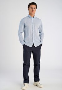 Giovane in piedi con le mani nelle tasche, indossa una camicia button-up azzurra chiara, pantaloni scuri e sneakers bianche su uno sfondo semplice.