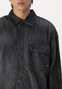 Camicia in denim nera con colletto, tasca frontale e chiusura a bottoni. Presenta una texture sbiadita e un piccolo logo giallo applicato.