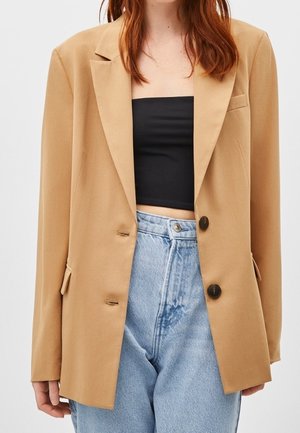 Femme portant un blazer oversize beige, un crop top noir sans bretelles et un jean bleu clair taille haute, montrée des épaules aux hanches.