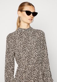 Vestido de estampa de leopardo com mangas longas e colarinho alto, apresentando uma cintura cinto e combinado com óculos de sol estilo gato-olho pretos.