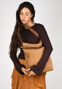 Bolso de hombro de ante marrón con un acabado texturizado, correa de cuero ajustable y detalle de hebilla metálica. Forma blanda y relajada con acentos mínimos.