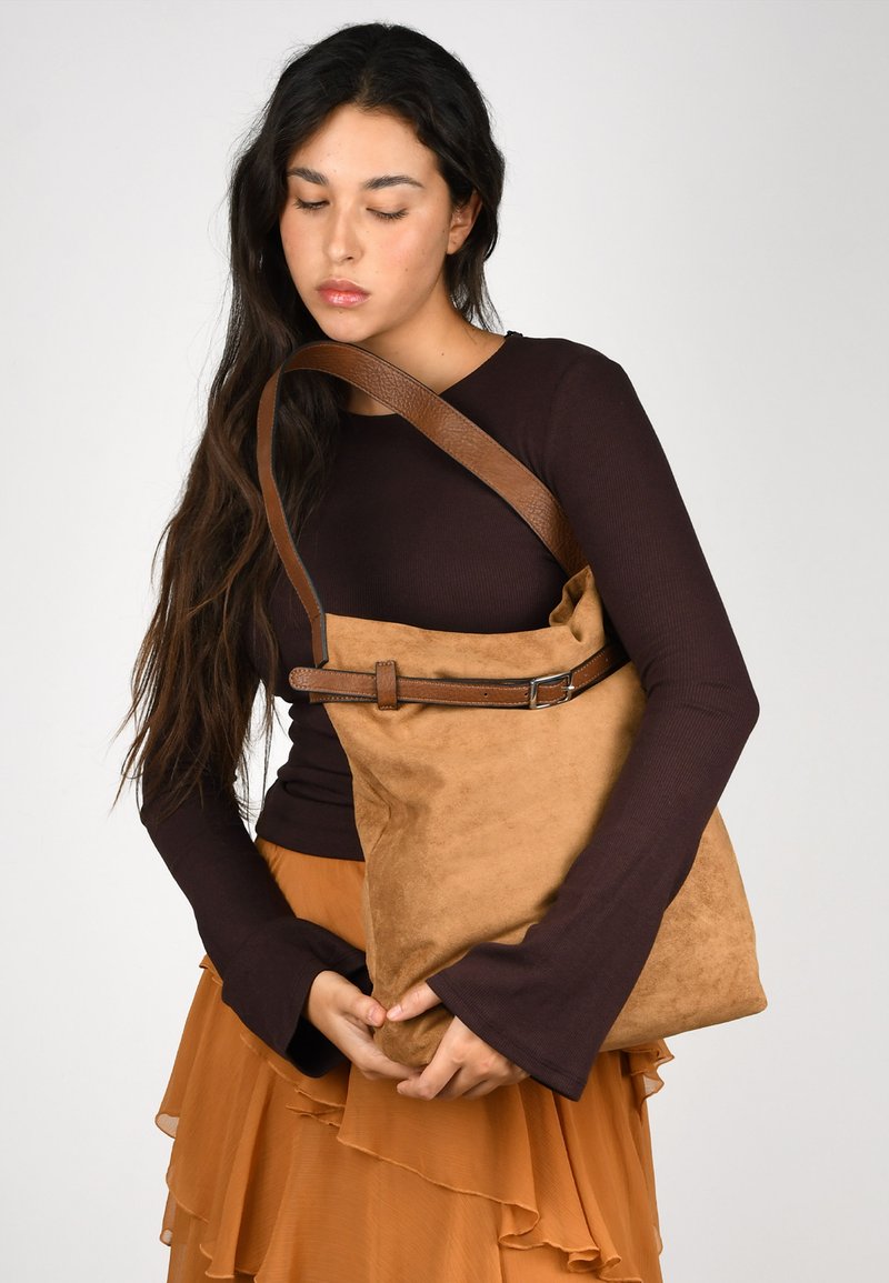 Bolso de hombro de ante marrón con un acabado texturizado, correa de cuero ajustable y detalle de hebilla metálica. Forma blanda y relajada con acentos mínimos.