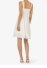 Robe blanche avec un corsage froncé, de larges bretelles et une jupe fluide. Portée avec des escarpins légers à brides. Texture lisse.